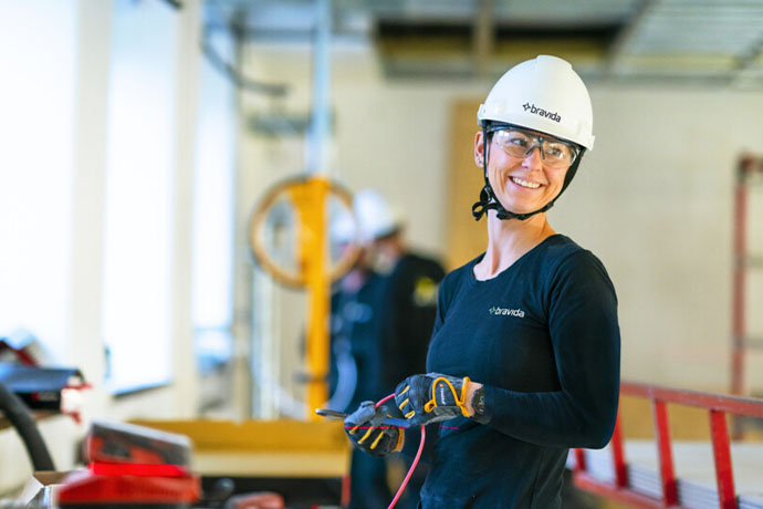 Person in work clothes with helmet and safety glasses holding glove in installation environment with equipment in the background.