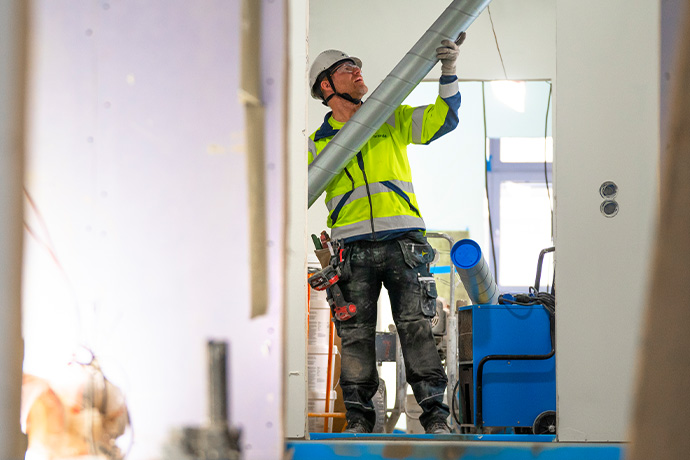 En ventilationstekniker som installerar ventilation på en byggarbetsplats. Personen är klädd i varseljacka, hjälm och arbetsbyxor, och håller i ett stort rör eller ventilationskanal som ska installeras. I bakgrunden syns olika byggmaterial och utrustning, inklusive en blå maskin och verktyg.