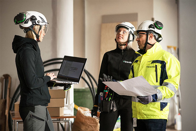 Tre personer i skyddshjälmar och arbetskläder diskuterar tekniska ritningar och information från en laptop på en byggarbetsplats.
