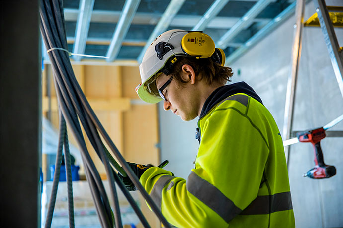 Elektriker i arbetskläder och skyddsutrustning arbetar med kablar inuti en byggnad, omgiven av byggnadsställningar och verktyg