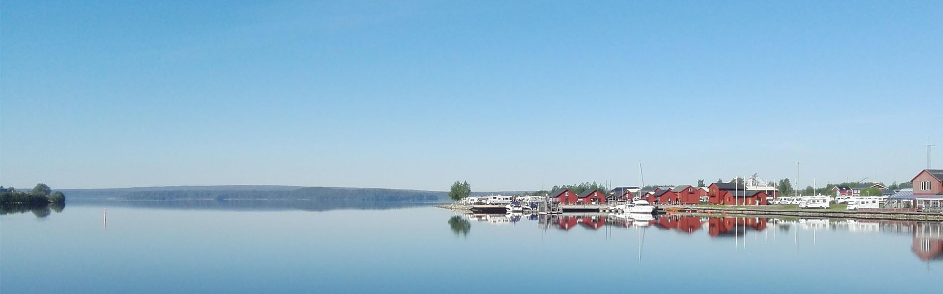 Bilden visar en stillsam hamnmiljö med spegelblankt vatten som reflekterar omgivningen. På vänster sida syns flera små båtar och segelbåtar förtöjda vid bryggor.
